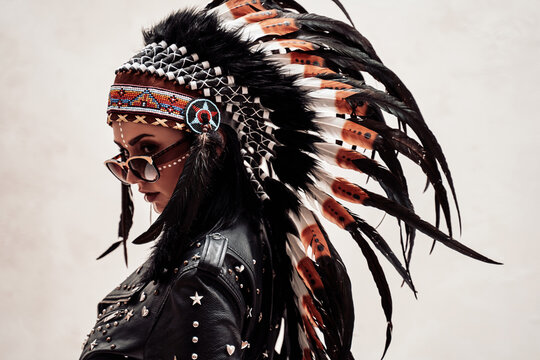 Assertive and cool young woman posing on a white background, wearing native american headdress, leather coat and sunglasses, looking dangerous with tribal make-up on