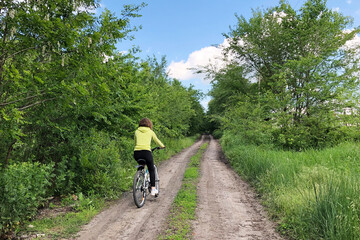 Obraz premium young female riding bicycle on countryside green road. Sport adventure in summer field. Healthy lifestyle concept.