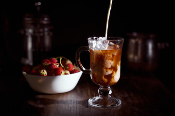 Ice coffee in a tall glass with cream poured over, ice cubes and beans on a old rustic wooden table with a plate of strawberries in the background.