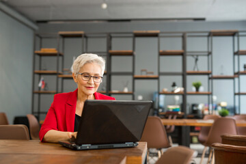 beautiful aged woman in a red jacket works in the office with a laptop