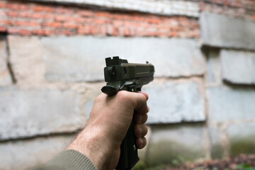 A gun in the hand of a Caucasian man against a brick wall of an abandoned building. Shooter with a gun of dark green.