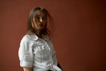 Fototapeta premium Serious confident young woman in rumpled white shirt posing against blank studio wall backgorund with copy space for your promotional information, her long hair falling on face because of wind