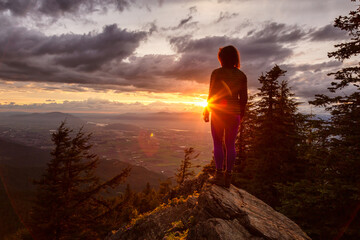 Fototapeta premium Adventurous Girl on top of a Rocky Mountain overlooking the beautiful Canadian Nature Landscape during a dramatic Sunset. Taken in Chilliwack, East of Vancouver, British Columbia, Canada.