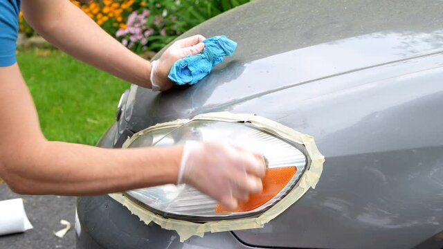 Man cleaning car headlights taped to protect paint rubbing with blue shop towel closeup on driveway of house outside
