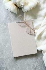 Flat lay composition with beige paper notebook, hipster glasses, textile cloth and peonies on concrete stone background. Elegant feminine workspace top view