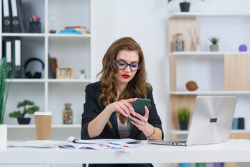 Smiling young business woman in stylish clothes sitting in cozy modern office and browsing something on smartphone.