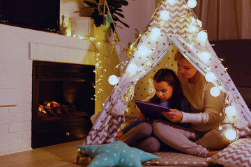 Mother and daughter are sitting in a teepee tent, reading stories with the flashlight. Happy family.