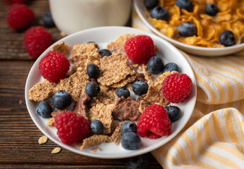Corn flakes with blueberries and raspberries in white bowl in dark wooden desk.