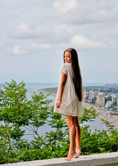 Portrait of young model girl in white dress. Sea at background. Fashion, pretty, barefoot teenage girl with long hair outdoors. Warm summer day and vacation time.