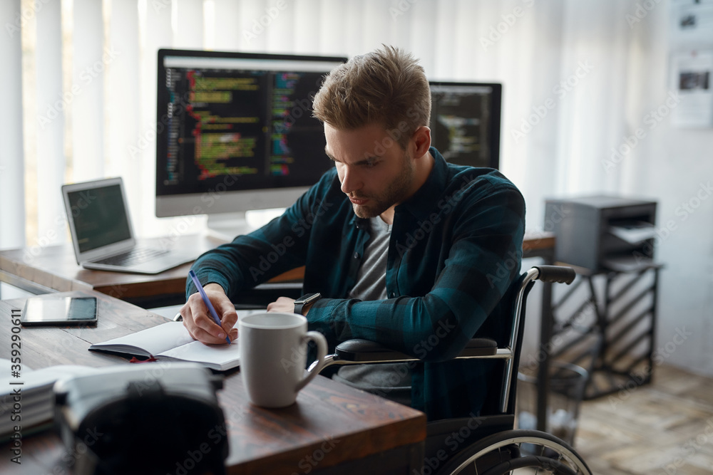 Wall mural side view of young concentrated male web developer in a wheelchair making some notes while sitting a