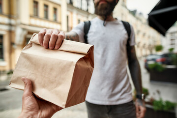Cropped shot of delivery man holding paper bag while giving away order to a customer. Courier,...
