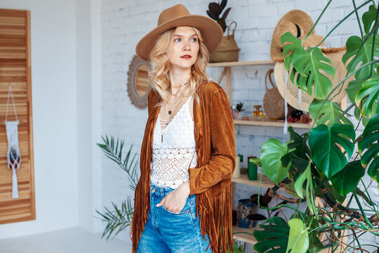 Fashionable lady wearing stylish boho outfit with hat, brown suede jacket, crochet top, many golden chains, jeans, posing in cozy interior with green  plants, white walls. Copy, empty space for text