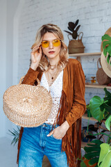 Fashionable woman wearing stylish boho outfit with yellow sunglasses, brown suede jacket, white crochet top, trendy round wicker straw bag, handbag, posing in cozy room © Victoria Fox