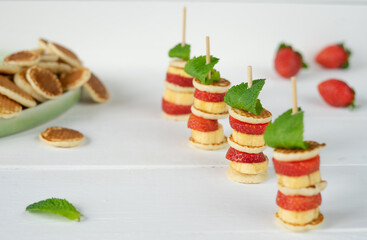 Four mini pancakes on a skewer with a mini banana, strawberries and fresh green mint. Fresh whole strawberries and a lot of mini pancakes in a green plate in the background. In the foreground is a lea