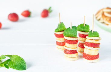 Four mini pancakes on a skewer with a mini banana, strawberries and fresh green mint. Fresh whole strawberries and a lot of mini pancakes in a green plate in the background. In the foreground is a lea