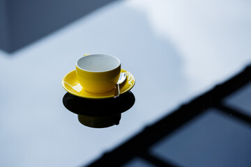 Yellow cup for coffee or tea with a saucer on a glass table.
