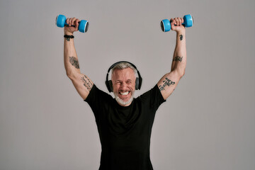 Fototapeta na wymiar Do it for life. Middle aged muscular man in black t shirt and headphones closing eyes while holding blue dumbbells, posing in studio over grey background