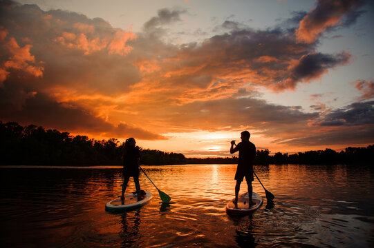 Rear View Of Two People On Sup Boards Which Floating On The River At Sunset