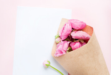 A bouquet of pink ranunculus flowers in craft wrapping paper. A white sheet of paper lies on pink paper. Top view, copy space. Concept Mother's Day, Family Day, Valentine's Day