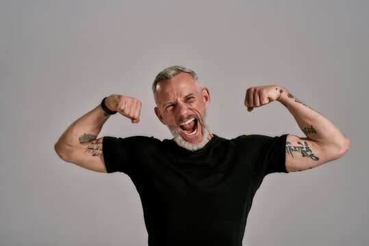 Be More, Do More. Angry Middle Aged Muscular Man In Black T Shirt Shouting At Camera, Showing His Biceps While Posing In Studio Over Grey Background