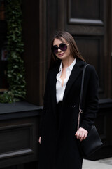 Obraz premium Outdoor photo of brunette lady walking on street background in cold autumn day. Fashion street style portrait. wearing dark casual trousers, white shirt, sunglasses and black coat. Fashion concept.