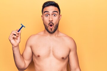 Young handsome man with beard shirtless holding depilation razor over yellow background scared and...