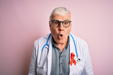 Senior handsome hoary doctor man wearing stethoscope and red HIV ribbon on coat afraid and shocked with surprise expression, fear and excited face.