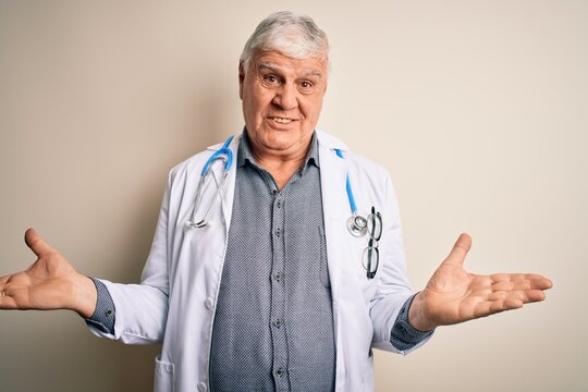 Senior handsome hoary doctor man wearing coat and stethoscope over white background clueless and confused expression with arms and hands raised. Doubt concept.
