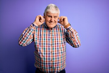 Senior handsome hoary man wearing casual colorful shirt over isolated purple background covering ears with fingers with annoyed expression for the noise of loud music. Deaf concept.