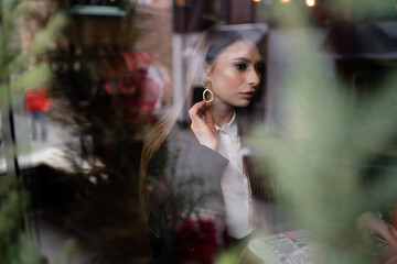 Beautiful brunette girl behind the glass, portrait of young woman in window, concept of female emotions. female model looking through the window. Winter cafe concept.