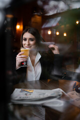Beautiful brunette girl behind the glass, drinking hot drink portrait of young woman in window, concept of female emotions. female model looking through the window. Winter cafe concept.