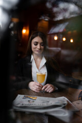 Beautiful brunette girl behind the glass, drinking hot drink portrait of young woman in window, concept of female emotions. female model looking through the window. Winter cafe concept.