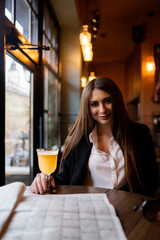 Beautiful woman with brunette hair in dark clothes looking at window and drinking orange hot drink. Concept of cozy places and cold winter. Concept of cafe and coffee shop. Cosy and atmosphere place.
