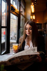 Beautiful woman with brunette hair in dark clothes looking at window and drinking orange hot drink. Concept of cozy places and cold winter. Concept of cafe and coffee shop. Cosy and atmosphere place.
