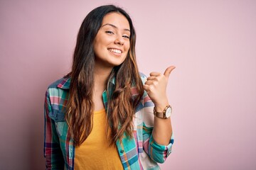 Young beautiful brunette woman wearing casual colorful shirt standing over pink background smiling with happy face looking and pointing to the side with thumb up.