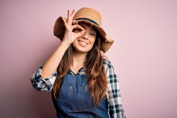 Young beautiful brunette farmer woman wearing apron and hat over pink background doing ok gesture with hand smiling, eye looking through fingers with happy face.