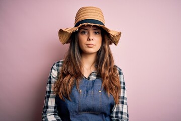 Young beautiful brunette farmer woman wearing apron and hat over pink background with serious expression on face. Simple and natural looking at the camera.