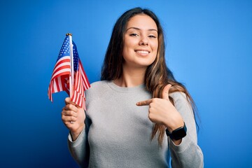 Young beautiful brunette patriotic woman holding american flag celebrating 4th of july with surprise face pointing finger to himself