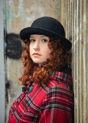Portrait of a young woman with curly red hair, wearing a red tartan coat, set in a rough urban setting.