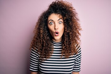 Young beautiful woman with curly hair and piercing wearing casual striped t-shirt afraid and...