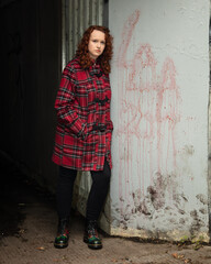 Portrait of a young woman with curly red hair, wearing a red tartan coat, set in a rough urban...