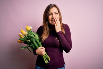 Young blonde woman holding romantic bouquet of yellow tulips flowers over pink background laughing and embarrassed giggle covering mouth with hands, gossip and scandal concept