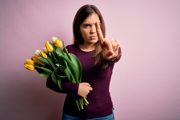 Young blonde woman holding romantic bouquet of yellow tulips flowers over pink background Pointing with finger up and angry expression, showing no gesture