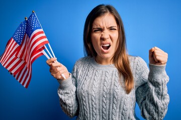 Young patriotic woman holding usa flag on independence day 4th of july over blue background annoyed and frustrated shouting with anger, crazy and yelling with raised hand, anger concept