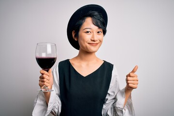 Young beautiful asian sommelier girl drinking glass of red wine over isolated white background happy with big smile doing ok sign, thumb up with fingers, excellent sign