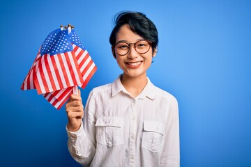 Young beautiful asian patriotic girl holding united states flags celebrating independence day with a happy face standing and smiling with a confident smile showing teeth