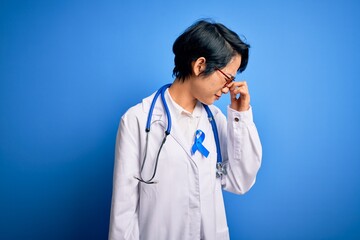 Fototapeta premium Young beautiful asian doctor girl wearing stethoscope and coat with blue cancer ribbon tired rubbing nose and eyes feeling fatigue and headache. Stress and frustration concept.