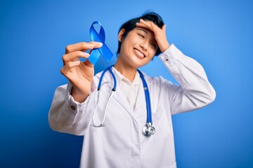 Young beautiful asian doctor girl wearing stethoscope and coat holding blue cancer ribbon stressed with hand on head, shocked with shame and surprise face, angry and frustrated. Fear and upset.