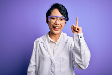 Young beautiful asian scientist girl wearing coat and glasses over purple background pointing finger up with successful idea. Exited and happy. Number one.