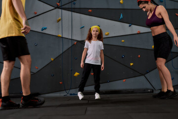 Fototapeta premium Active woman and little girl going to climb, male instructor helping them to warm up against artificial training climbing wall. Concept of sport life and rock climbing